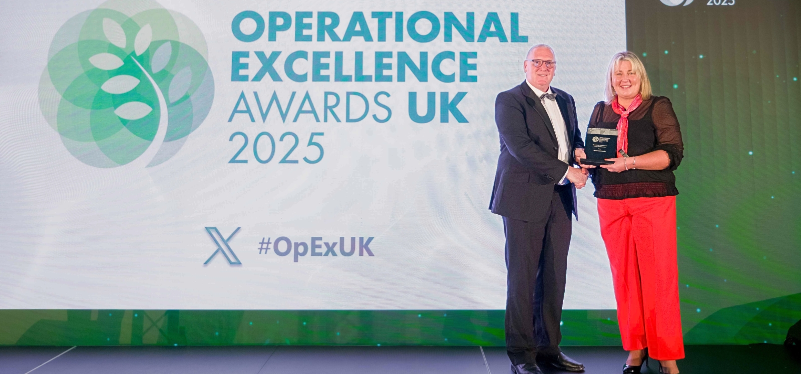 Woman in bright red trousers accepting an award trophy from a besuited man at an awards ceremony. Background presentation screen reads 'Operational Excellence Awards UK 2025 / #OpExUK'.